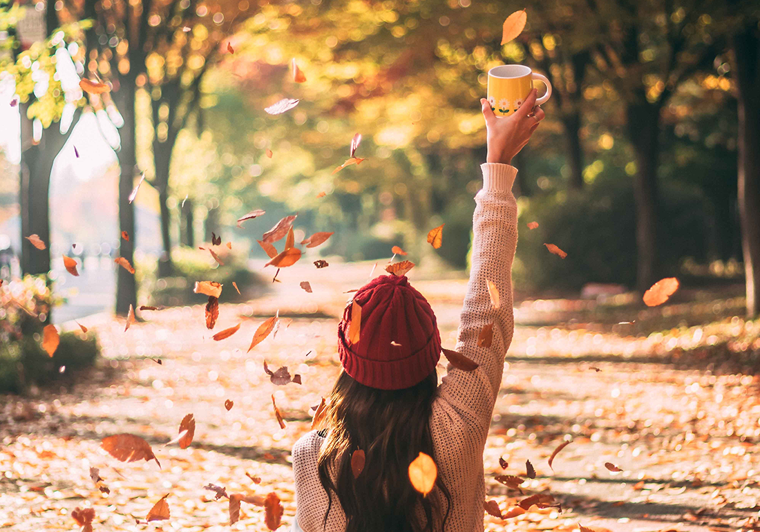 Junge Frau mit Pullover und roter Mütze, die mit erhobener Hand, die eine Tasse hält. auf einer Allee sitzt mit herabfallendem Herbstlaub