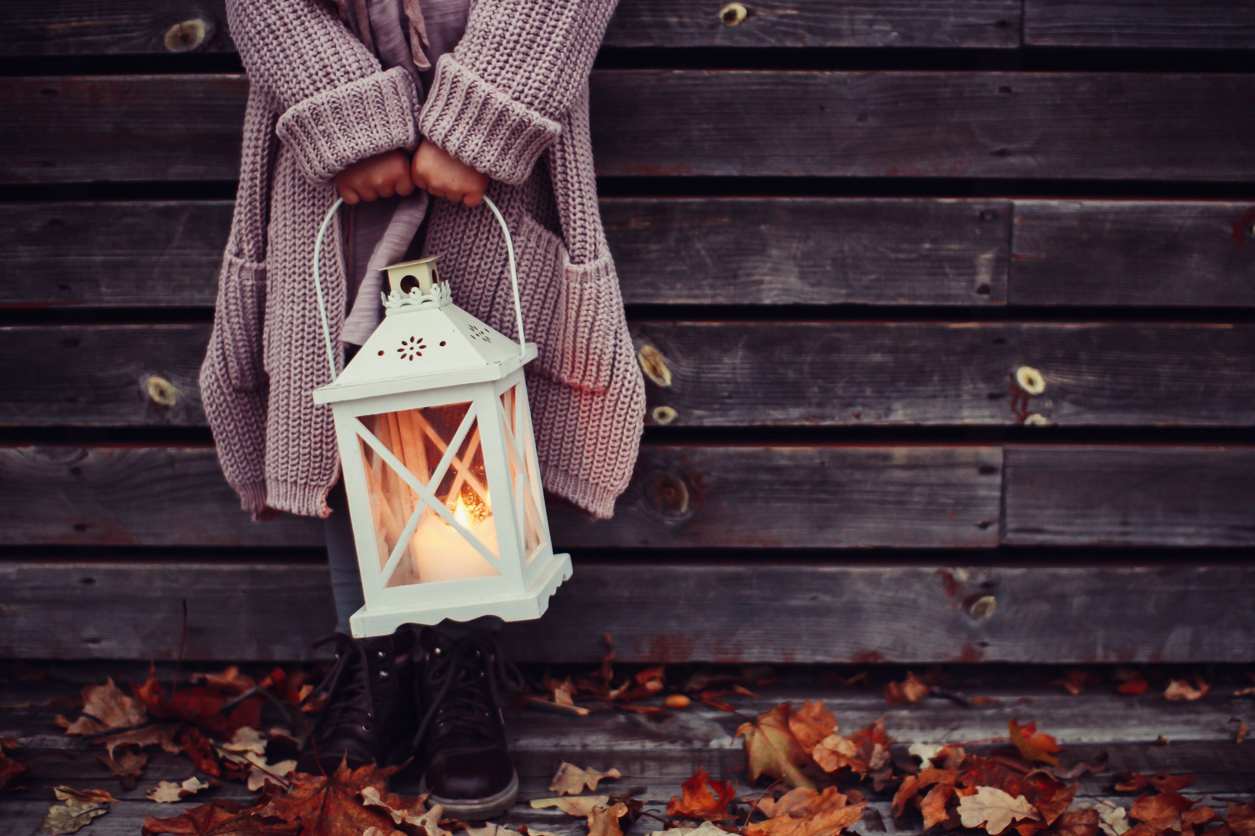 Kind das eine weiße Laterne hält und auf Herbstlaub steht. Lehnt sich gegen eine alte Holzwand