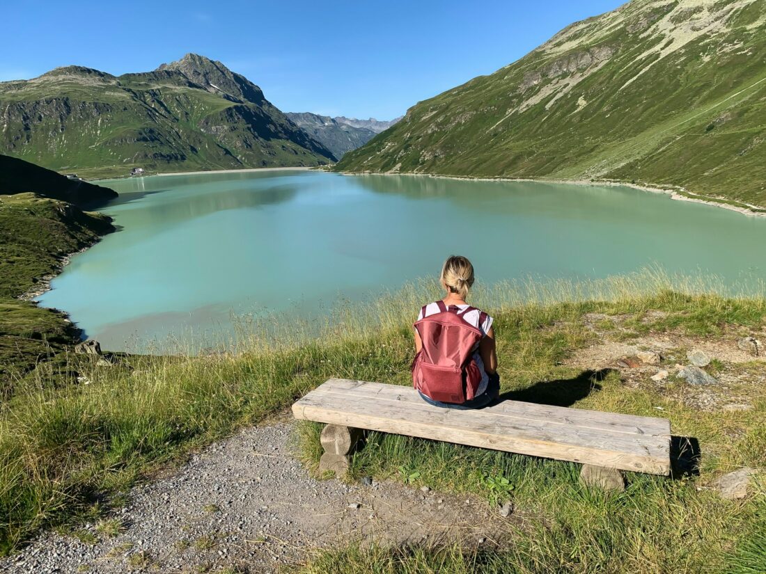 Alles über Vitamin D Beitragsbild: Frau die einen Rucksack auf dem Rücken hat und bei Sonnenschein auf einer Bank vor einem Bergesee sitzt.