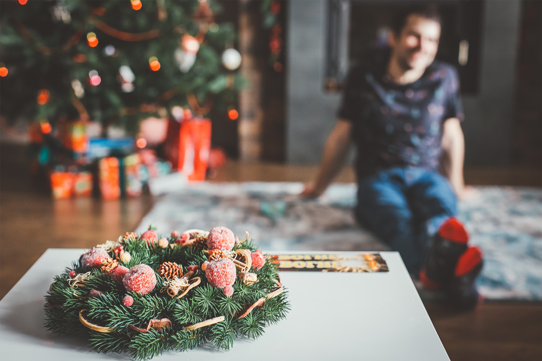 Weihnachtlicher Kranz mit Tannennadeln und ein entspannter Mann vor dem Weihnachtsbaum mit Geschenken sitzend. Stress in der Weihnachtszeit vermeiden